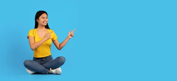 黄色のTシャツの中でかなり若い中国の女性の笑顔の床に座って 空の空間に指で青の背景 スタジオ パノラマに隔離されたポイント 素晴らしい広告 販売とオファー — ストック写真