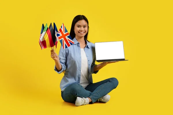 Cheerful glad millennial asian lady teacher, tutor with many different flags sits on floor, shows laptop with empty screen isolated on yellow background. Study and education online, learn language