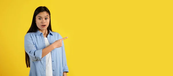 Glad surprised funny young japanese female with open mouth, pointing finger at empty space isolated on yellow background, panorama, studio. Shocking ad and offer, amazed facial expression and emotions