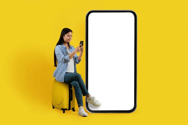 Glad young chinese woman sit on suitcase near huge phone with blank screen, typing on smartphone isolated on yellow background, studio. Travel at vacation, booking, online chat, blog, app and ad
