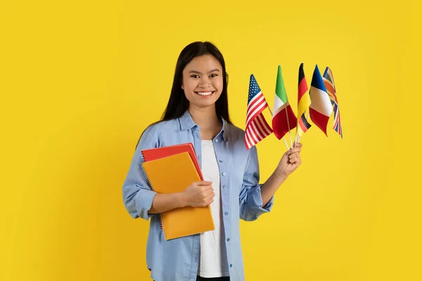 Satisfied young asian lady student or teacher with many international flags, notebooks looks at camera, isolated on yellow background, studio. Modern study and education, learn new language at school