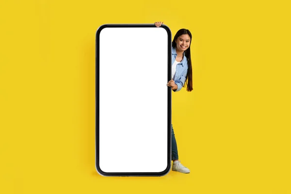 Glad funny young korean female peeking out from behind huge phone with blank screen isolated on yellow background, studio. Social networks, blog, app, ad and offer, modern tech for chat and study