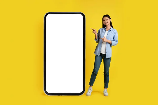 Social networks and chat. Cheerful happy young asian lady pointing finger at huge phone with blank screen for ad, isolated over yellow background, studio. New blog, app, offer and modern technology