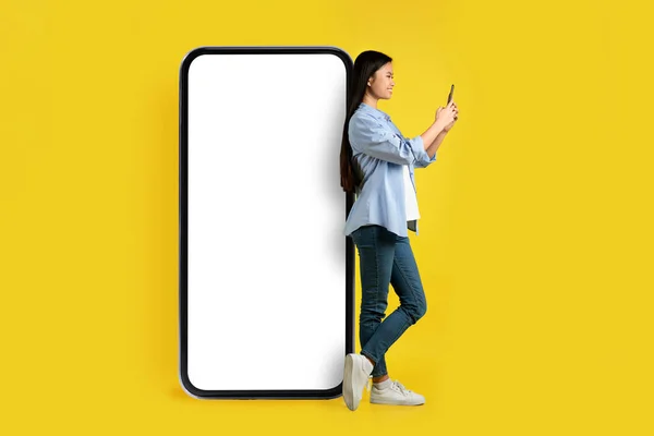 笑着拿着智能手机的年轻亚洲女人站在巨大的手机旁边 屏幕空白 背景是黄色的 工作室 社交网络 提供和现代技术中的在线聊天 — 图库照片
