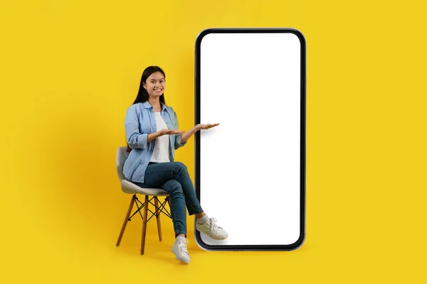 Cheerful young asian woman sits on chair, shows hands at huge phone with empty screen on yellow background, studio. Presentation, online chat in social networks, blog, app, ad, offer and modern device