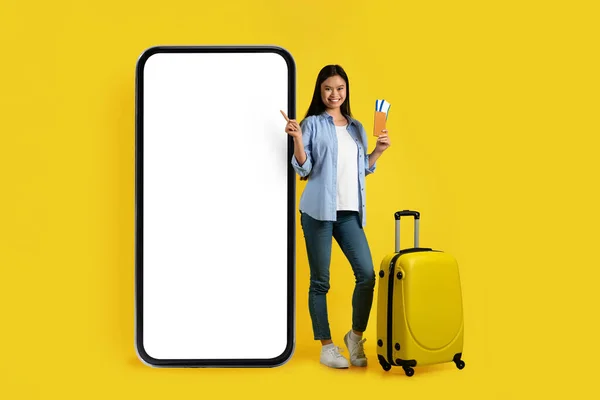 Glad young asian lady with suitcase, passport and ticket point finger at huge phone with empty screen, typing on smartphone isolated on yellow background, studio. Booking, online map, blog, app, ad