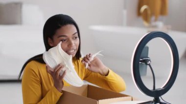 Moda blogu. Genç, Afro-Amerikan kadın etkileyicisi, yeni beyaz ayakkabıları inceliyor, akıllı telefondan evde yayın yapıyor, portreyi kapatıyor.