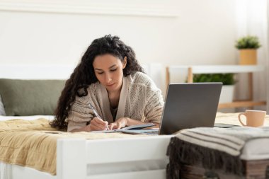 Bilgisayarlı genç bir kadın bilgisayarını kullanıyor, çalışıyor ya da online çalışıyor, iş toplantısı ya da webinar sırasında not alıyor, uzaktan ders alıyor, evde yatakta yatıyor. Mesafe çalışması, eğitim kavramı