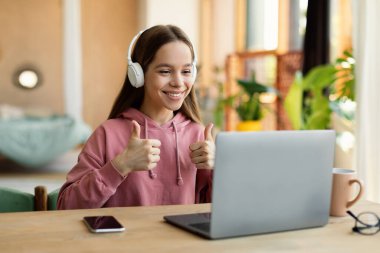 Kulaklıklı, evde dizüstü bilgisayarla çalışırken baş parmağını gösteren beyaz bir kız. Harika online ders, özel ders, uzaktan eğitim konsepti.
