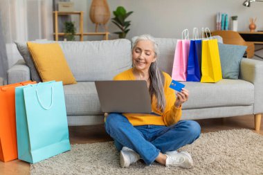 Alışveriş yaptıktan sonra beyaz saçlı, beyaz saçlı mutlu bir beyaz kadın oda içinde online alışveriş yapmak için kredi kartı ve laptop kullanıyor. Emekli alışverişkolik, bankacılık ve satış, covid-19