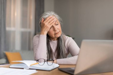 Depresyonda olan Avrupalı, gri saçlı olgun bir kadın, baş ağrısı çekiyor, stres yaşıyor ve odanın içinde mutsuz bir yüz ifadesi var. Sağlık sorunları, migren, covid-19 salgını sırasında evde aşırı çalışma ağrısı.