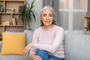 Neşeli, kendine güvenen, sakin, gri saçlı, Avrupalı, yaşlı bir kadın, kanepede oturuyor, boş zamanlarının tadını çıkarıyor ve oturma odasının iç tarafındaki kameraya bakıyor. Rahat evde mola, dinlenme ve rahatlama, boşluğu kopyala