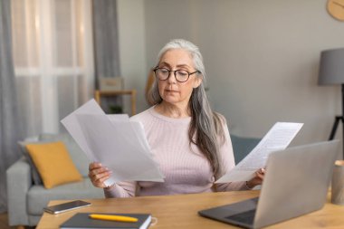 Gözlüklü, gri saçlı, ciddi bir Avrupalı kadın bilgisayarda ve odanın içindeki belgelerde çalışıyor. Muhasebe, fatura ödeme, vergi ödeme ve yeni normal, eğitim ve iş uzaktan evde, covid-19