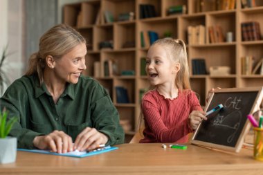 Dost canlısı çocuk geliştirme uzmanı bir kadın çocuğun önünde oturuyor ve öğrenciye kelimeleri öğretiyor, kız tahtaya kediler yazıyor.