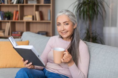 Gri saçlı, neşeli Avrupalı son sınıf öğrencisi kadın kahve içiyor ve oda içinde boş zamanlarında en sevdiği kitabı okuyor. Konforlu evde hobiler yalnız dinlenme ve içkiyle rahatlama zamanı covid-19 salgını
