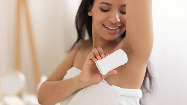 Happy Black Lady Applying Antiperspirant On Armpits For Freshness And Sweat Protection Standing Wrapped In Towel In Modern Bathroom At Home. Bodycare Cosmetics Concept. Panorama, Selective Focus