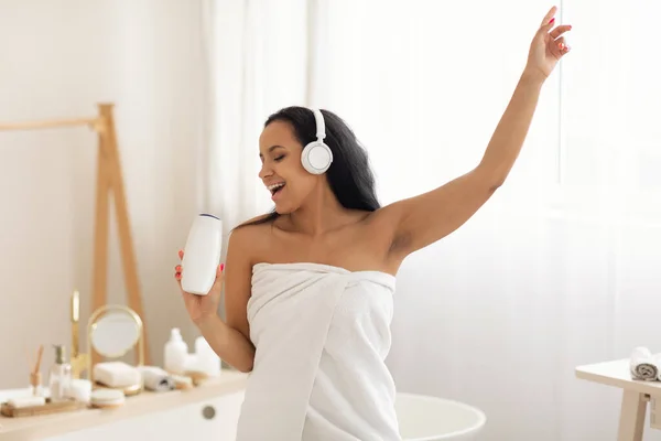 Happy Black Female Singing Holding Shampoo Bottle Like Microphone Enjoying Beauty Routine And Advertising Cosmetics Standing Wrapped In Towel In Modern Bathroom At Home, Wearing Headphones
