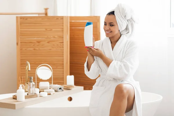 シャンプーボトルを見せる魅力的な黒人女性現代的な浴室の屋内で白いバスローブを着て浴槽に座って化粧品製品をポーズをお勧めします 女性の広告シャワージェル — ストック写真