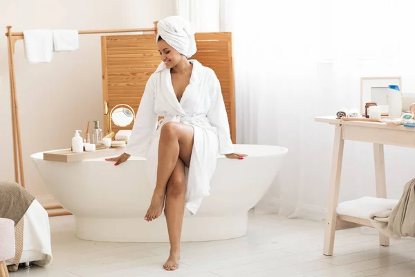 在家里的现代浴室里 穿着白色浴衣的女人坐在浴缸里 用化妆品护理身体和皮肤 美容美发 健康与温泉的概念 — 图库照片