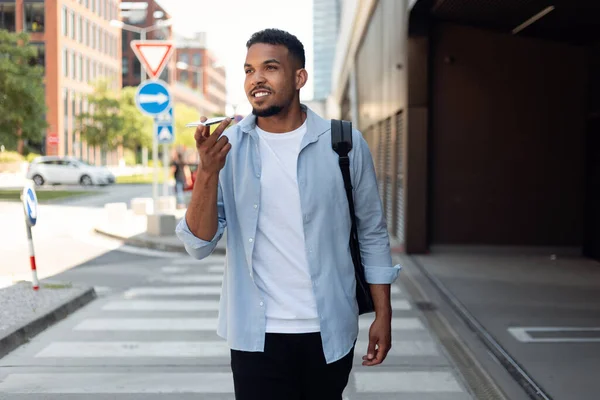 Handsome african american guy recording audio message on mobile phone, having conversation with his friend while walking in the city. Modern technologies and communication concept