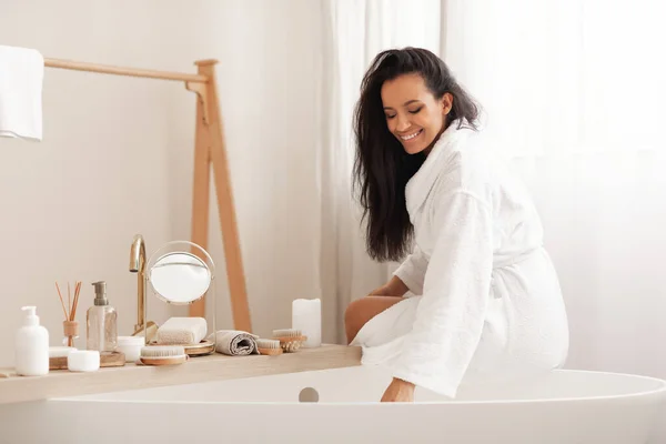 Beauty Routine. Happy Mixed Woman Sitting On Bathtub Preparing Bath Adding Sea Salt In Modern Bathroom At Home. Bodycare Cosmetics, Wellness And Spa Concept