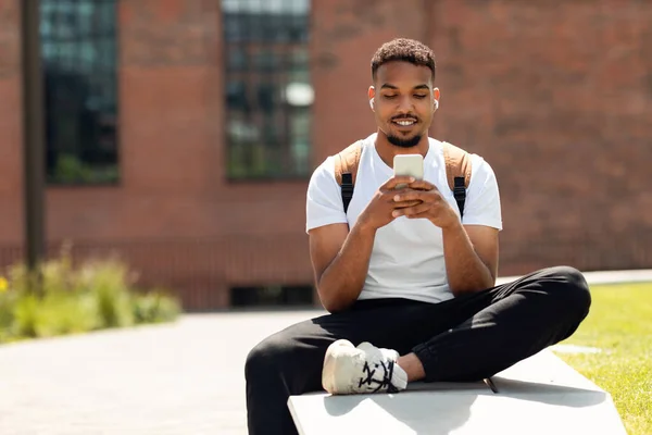 Modny Czarny Student Korzystający Smartfona Sms Muzyki Odsłuchowej Słuchawkach Siedzący — Zdjęcie stockowe