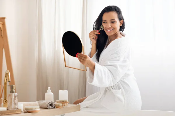 Cheerful Mixed Woman Massaging Face Stone Roller Holding Mirror Sitting — Stock Photo, Image