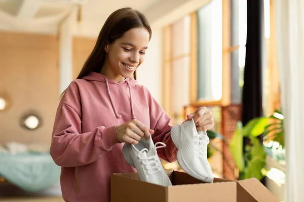 Happy teen girl buyer holding new footwear unpacking cardboard box in bedroom interior at home. Young customer receiving shoes after successful online shopping