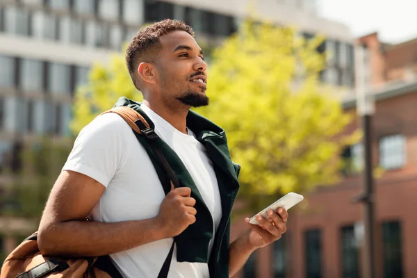 Handsome african american college guy using smartphone outdoors, walking in urban area after classes, texting online on cellphone. Black male student holding mobile phone. People and gadgets