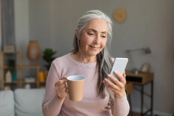 Smiling european old woman with gray hair looks at smartphone, have chat and drinks coffee in room interior. News and new app, offer and advertisement, surfing in internet during covid-19 pandemic