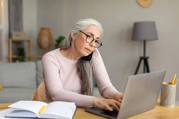 Serious european old lady with gray hair in glasses works on computer and calls by phone in room interior, copy space. Meeting remotely and new normal, study and business at home during covid-19 virus