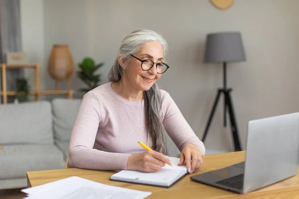 满脸笑容的戴眼镜灰白头发的欧洲老太太看着电脑 在房间里的桌子边做笔记 流行性感冒爆发期间 在家与家人进行远程和新的正常接触 工作和经商 — 图库照片