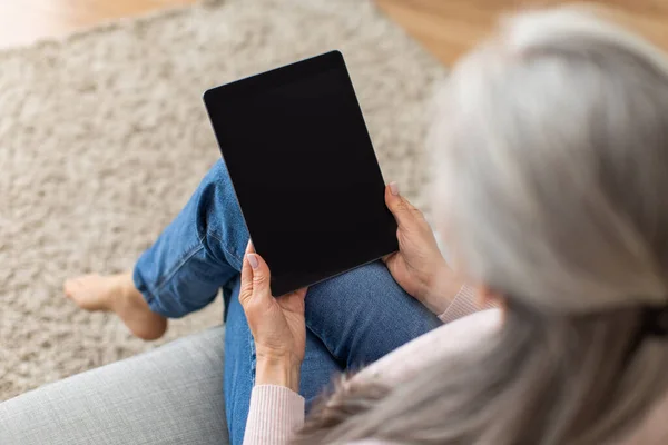 European old lady with gray hair looks at tablet with empty screen, surfing in internet in room interior, over shoulder view. Social distance, new app, offer and ad, chat at home due covid-19 pandemic
