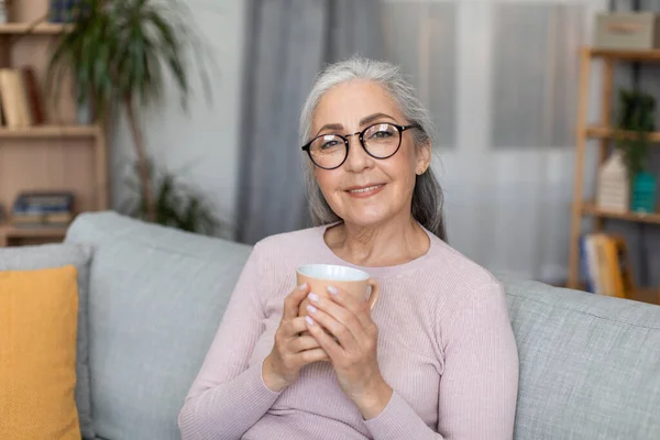 这位有着一头灰白头发 头戴眼镜的快乐镇定的欧洲老妇人的画像 拿着杯子喝酒 在客厅里享受着闲暇时光 喝咖啡休息 在家休息和放松以及生活方式 — 图库照片