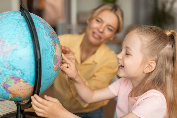 Cheerful female tutor teaching child geography, girl learning about the world countries during tutoring class at home, pointing at globe and smiling