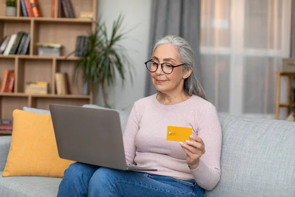 Smiling european senior woman with gray hair in glasses works on laptop and shows credit card in living room interior. Online bill payment, shopping, sale and order purchase at home during covid-19
