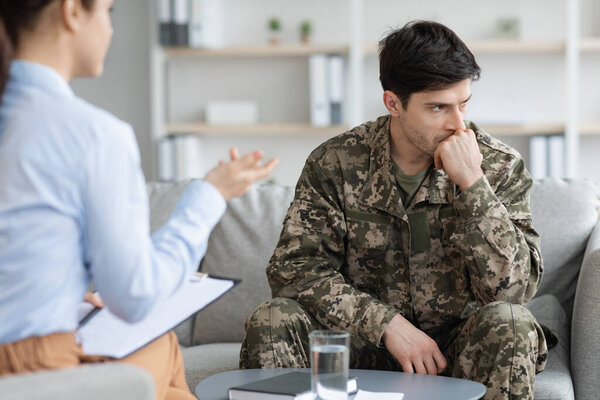 Pensive young man in military uniform veteran sitting on couch, touching his mouth and looking aside while having conversation with female doctor, soldier attending psychotherapist at clinic