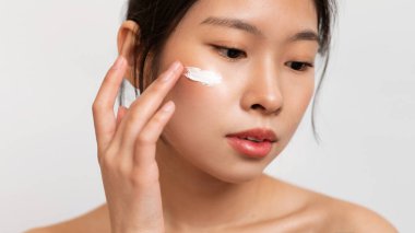 Güzel Asyalı bir bayanın yaşlanma karşıtı ya da nemlendirici yüz kremini yanağına sürmesi, beyaz stüdyo arka planında durması, panorama. Yüz bakımı, kaldırma, besleyici kozmetik konsepti