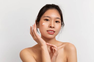 Genç Koreli kadın yüzüne krem sürüyor, cildini nemlendiriyor ve kameraya bakıyor, beyaz stüdyo arka planında poz veriyor. Yüz derisi kozmetik konsepti