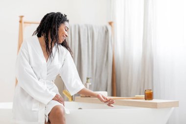 Ev Kaplıcası. Küvette oturan ve köpüğe dokunan güzel siyah kadın beyaz ipek bornoz giyen çekici Afro-Amerikan kadın banyo yapmaya hazırlanıyor evdeki banyoda dinleniyor ve boşluğu kopyalıyor.