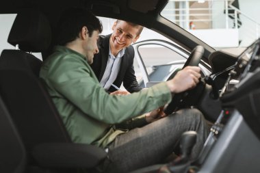 Yeni araba alan mutlu bir milenyum adamı satıcılarla araba satın alma detaylarını açıklığa kavuşturuyor galeride oto galerisinde oturuyor. Genç beyaz adam galeride otomobil satın alıyor.