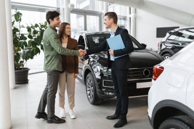 Genç beyaz aile anlaşma yapıyor, modern otomobil alıyor ya da kiralıyor, galeride araba satıcısıyla tokalaşıyor. Milenyum çifti otomobil galerisinden yeni bir araç satın alıyor