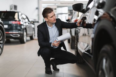 Yakışıklı genç satış müdürü lüks araba salonunda yeni otomobil kontrolü yapıyor, tam boy. Modern otomotiv bayiliğinde milenyum sigorta acentesi çalışıyor