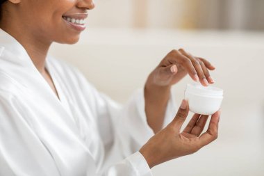Smiling black woman holding open jar with moisturising cream, unrecognizable african american female wearing white silk robe making beauty treatments at home, enjoying skin care routine, cropped shot clipart