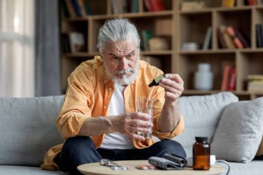 Hipertansiyondan muzdarip hasta sakallı yaşlı bir adam oturma odasında koltukta oturuyor, ilaç kullanıyor, su bardağına damla damlatıyor, fotokopi çekiyor. Tansiyon ilaçları konsepti
