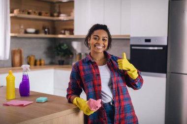 Binlerce yıllık neşeli Afro-Amerikan kadın lastik eldivenli, paçavra ve temizlik malzemeleriyle modern mutfak içinde baş parmağını gösteriyor. Evde temizlik, ev işi, hijyen ve ev kadınlarını onayla