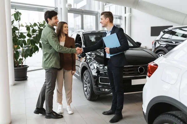 Young Caucasian family making deal, buying or renting modern automobile, shaking hands with car salesman at auto showroom. Millennial couple purchasing new vehicle at auto dealership