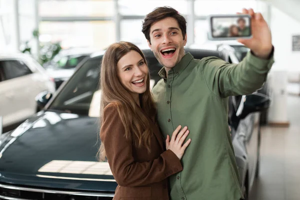 Excited young Caucasian couple taking selfie or filming video on smartphone near new auto at dealership. Cheerful millennial family making photo next to luxury vehicle at modern showroom