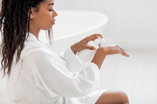 身体滋养 漂亮的非洲裔美国女人手握润肤乳液 身穿白色丝袍的迷人黑人女士在现代浴室的室内进行恐怖治疗 — 图库照片