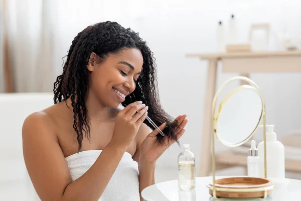 快乐的黑人女士在淋浴后被损坏的头发上涂上润肤油 在浴室内笑着坐在镜子旁边的非洲裔美国女人 在家里做日常的理发 穿上衣服 — 图库照片